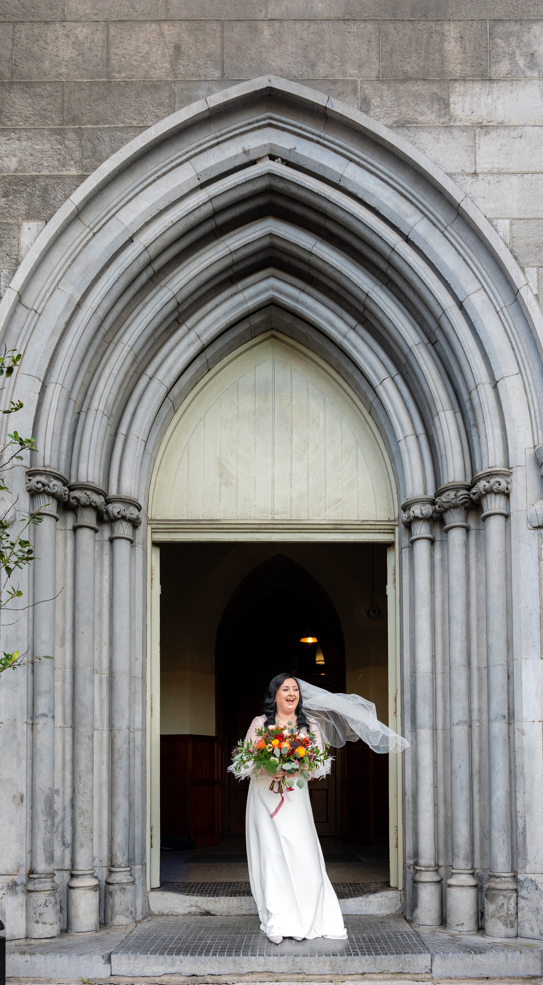 Noiva na porta da igreja com o véu levado pelo vento, casamento de C & S