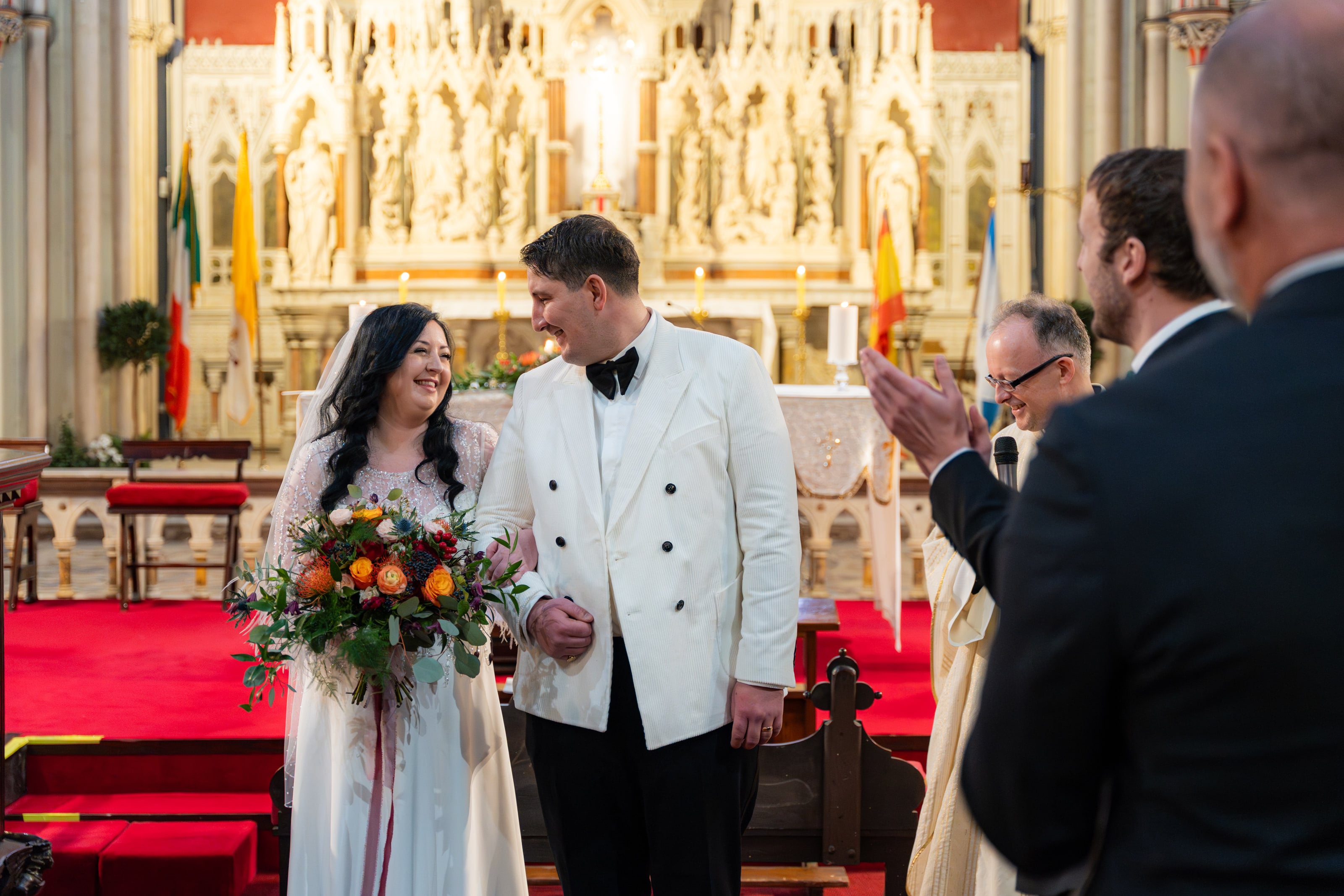 Noiva e noivo de paletó branco sorrindo diante do altar dourado, casamento de C & S