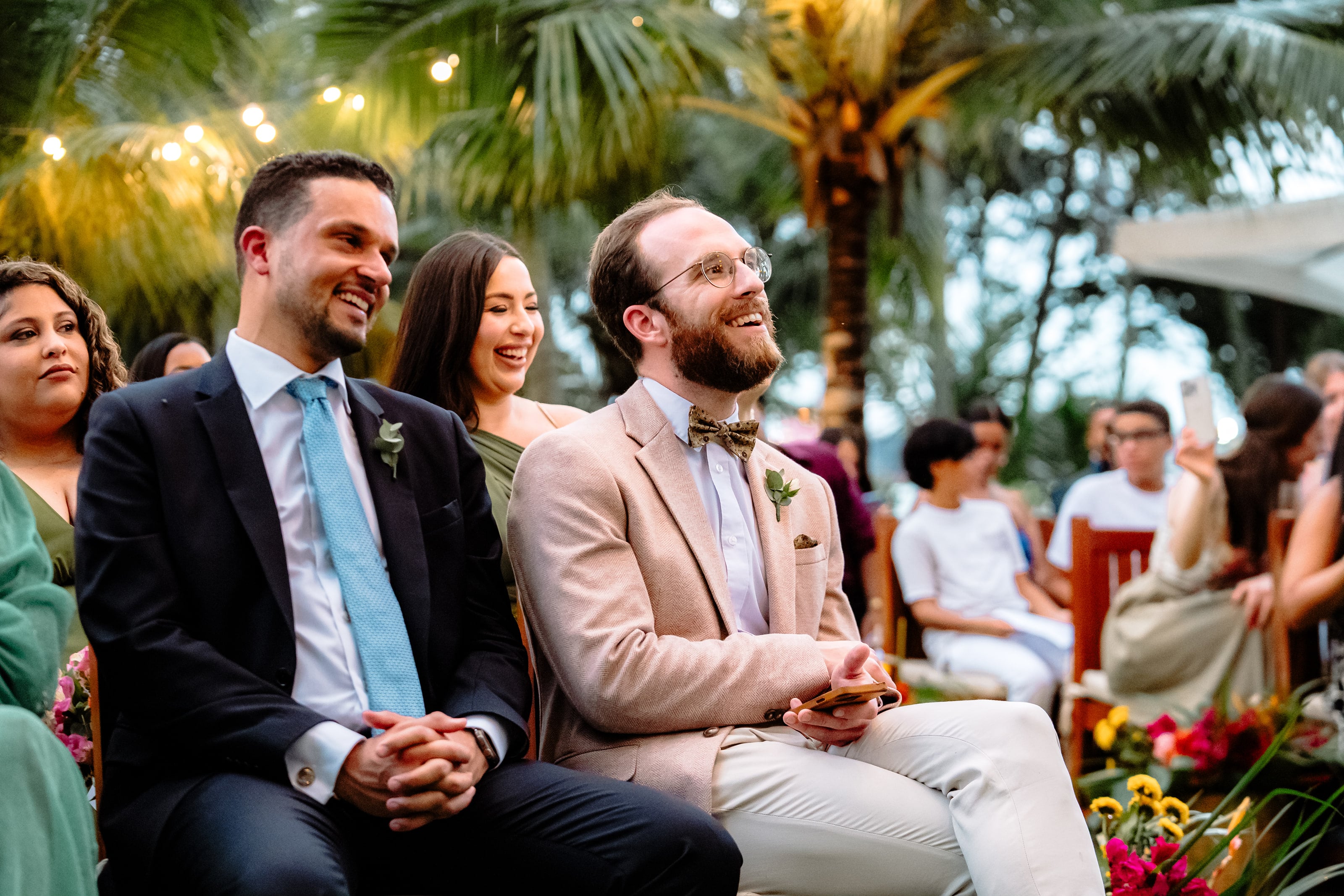 Convidados rindo sob as luzes entre os coqueiros, casamento de J & K