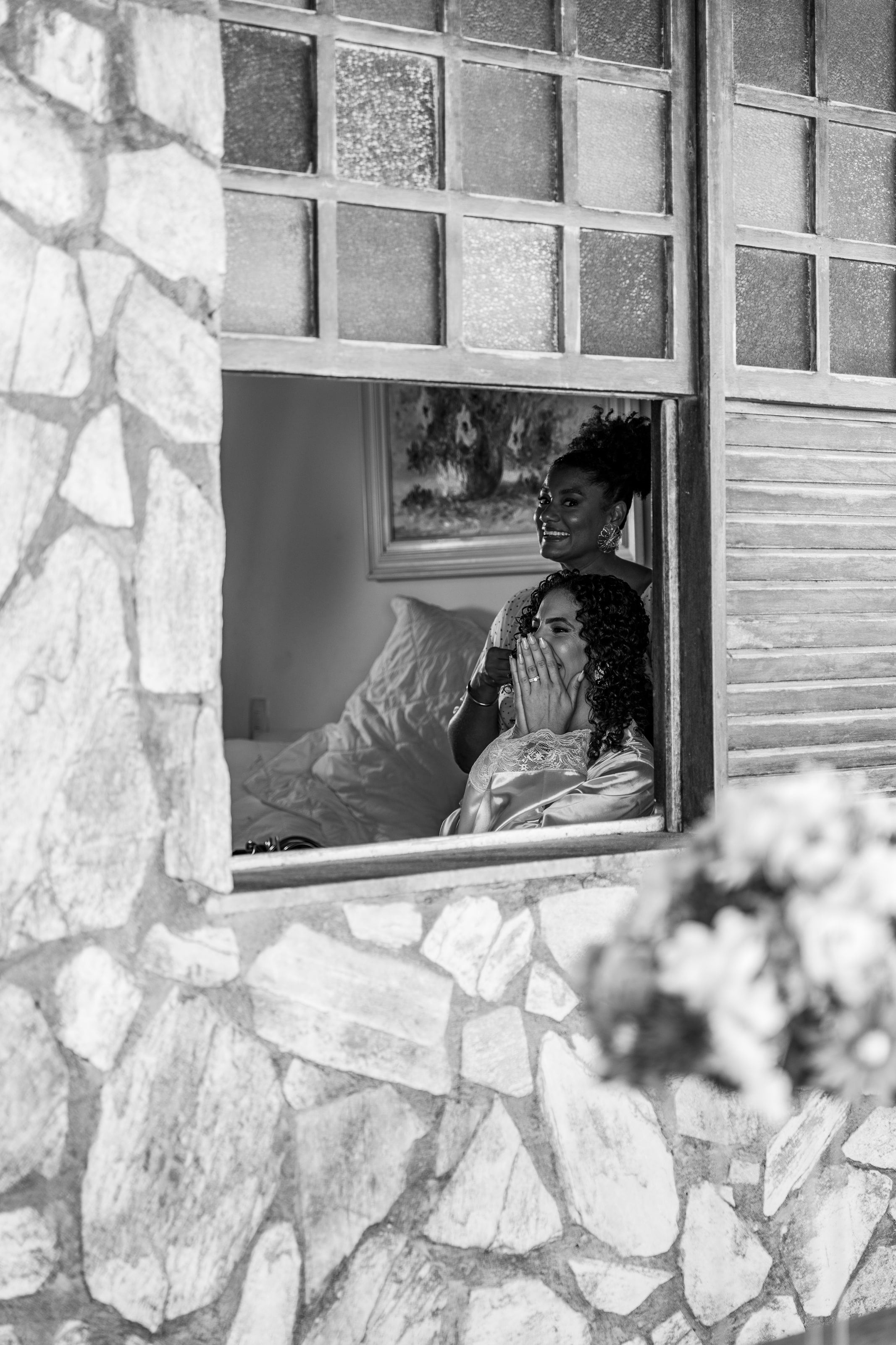 Noiva e uma amiga vistas pela janela de pedra durante a preparação, em preto e branco, casamento de L & M em Salvador