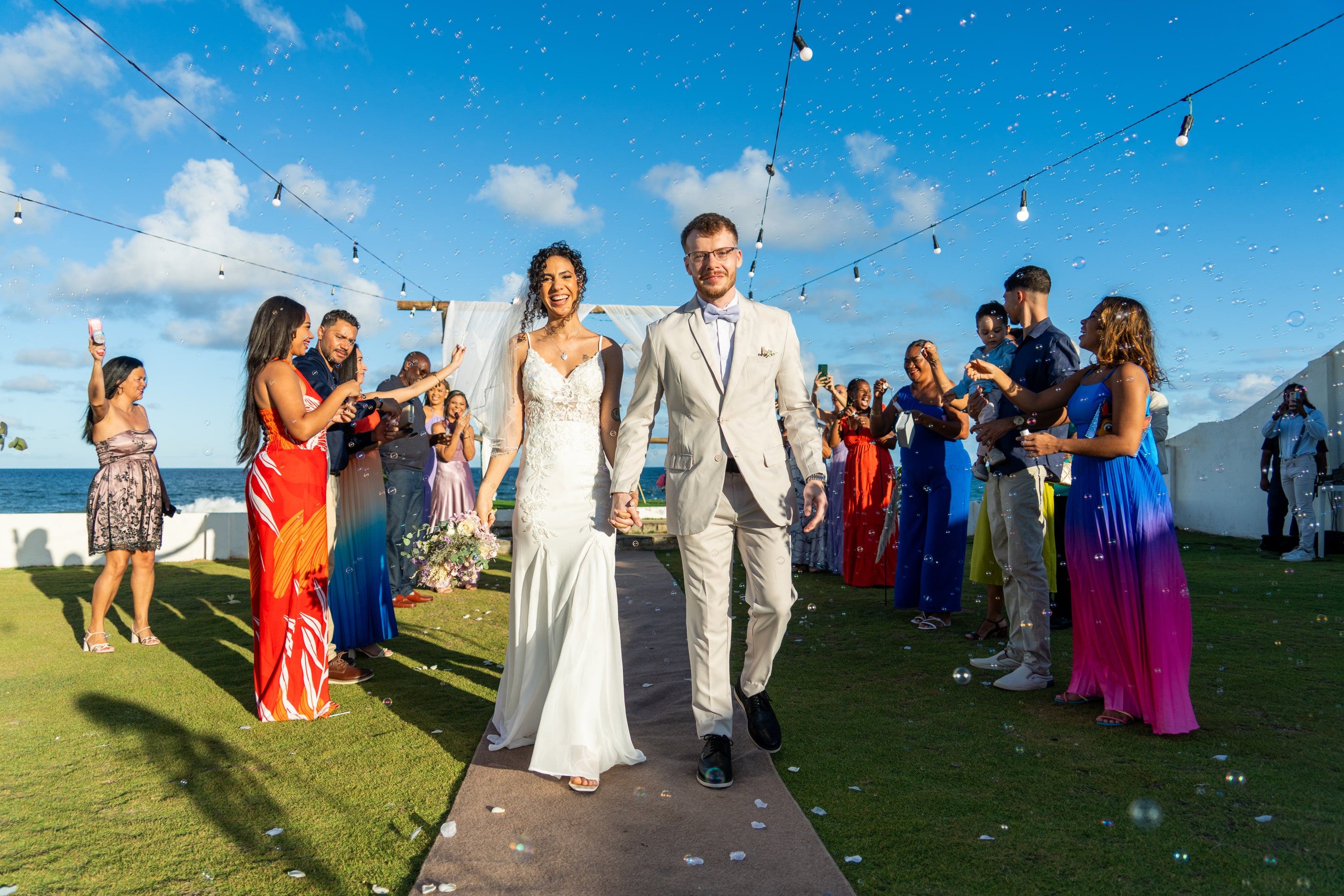 Casal saindo pelo corredor entre convidados de vestidos coloridos, casamento de L & M em Salvador