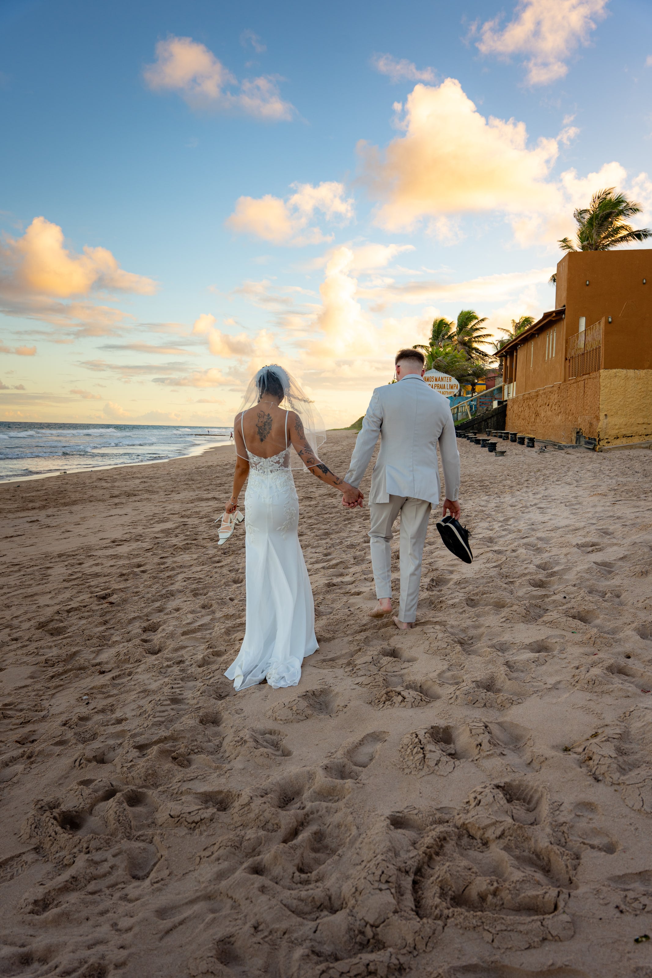 Casal caminhando descalço na praia, sapatos na mão, casamento de L & M em Salvador