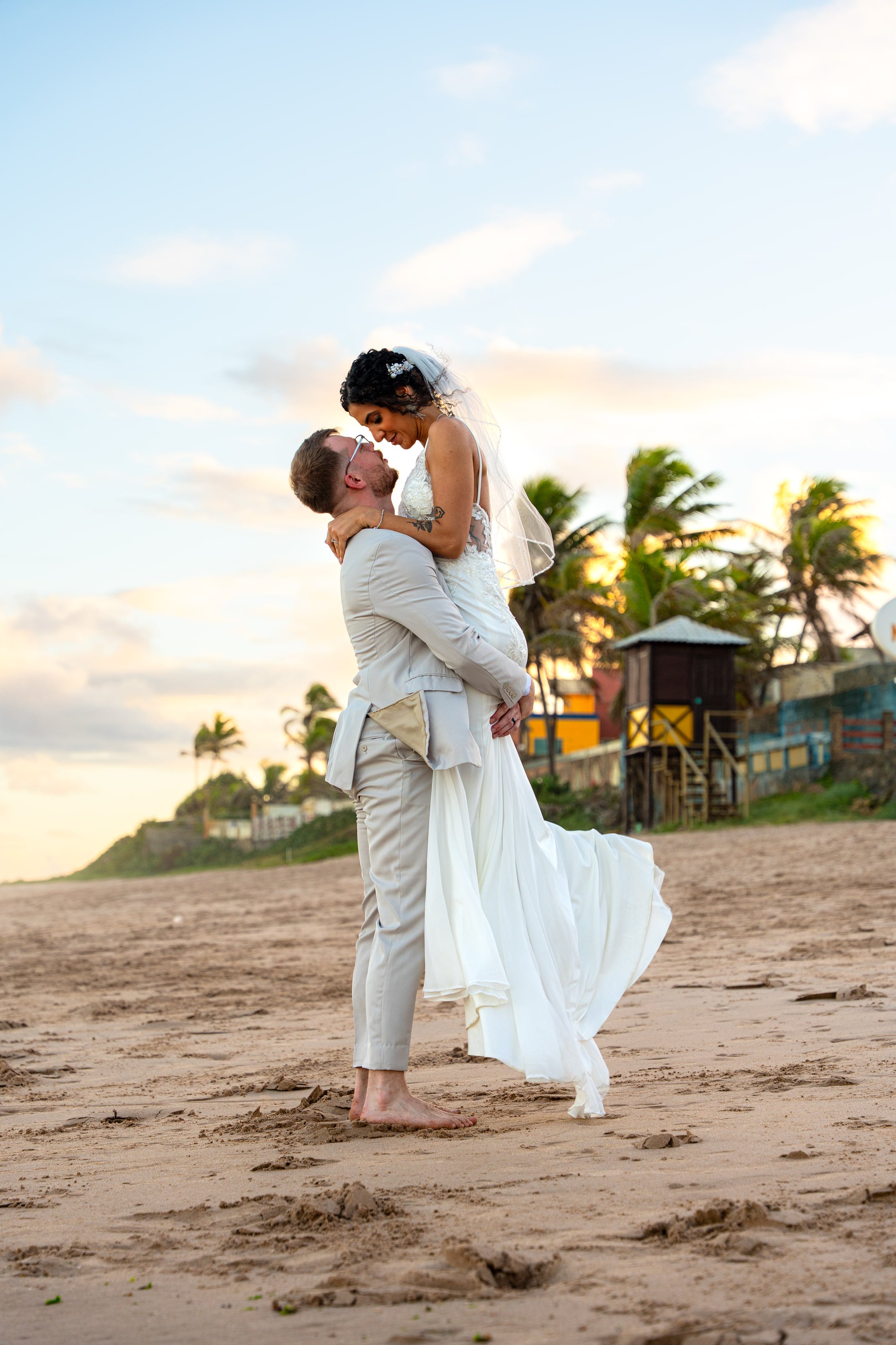 Noivo levanta a noiva na areia ao fim da tarde, com coqueiros ao fundo, casamento de L & M em Salvador