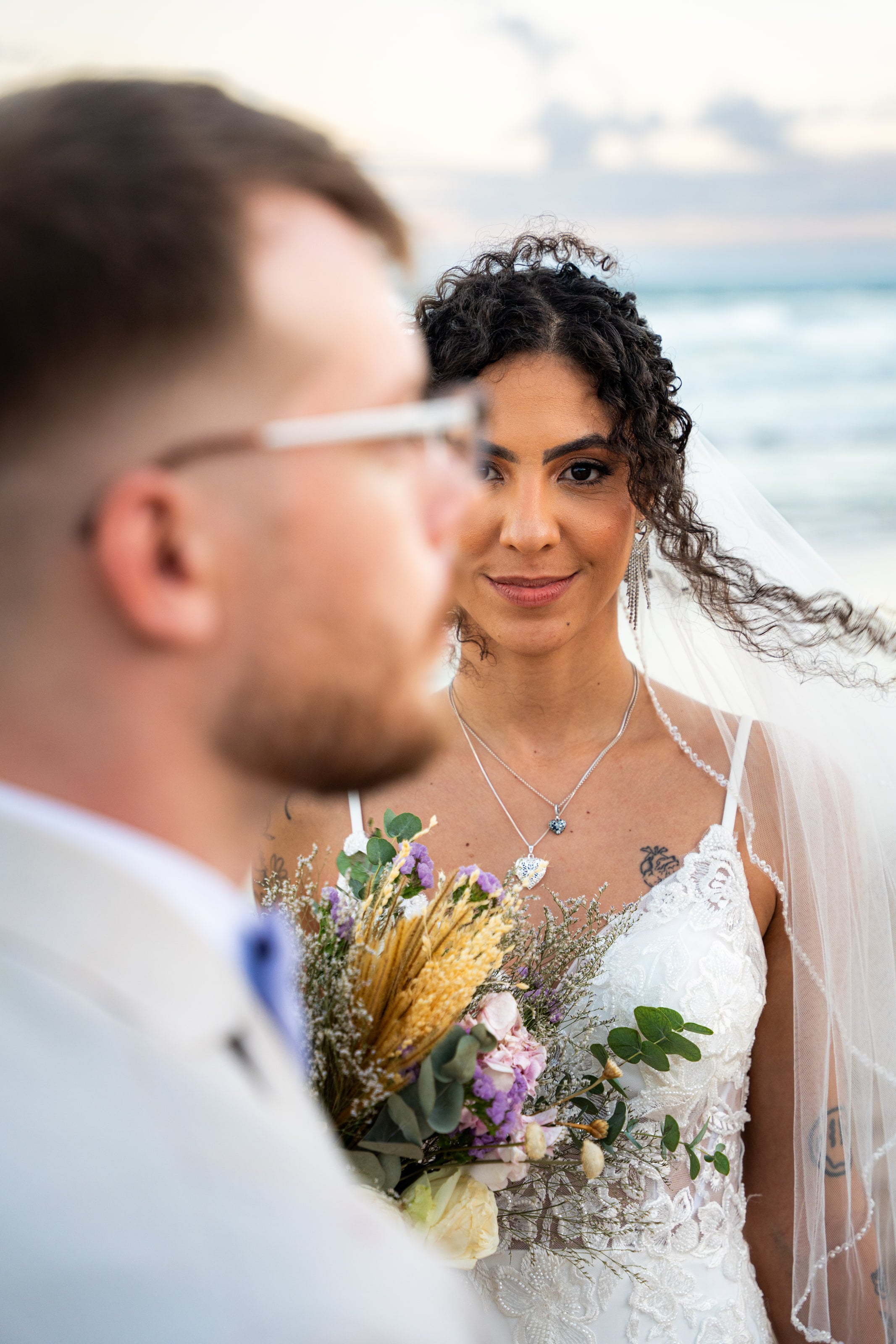Noiva olha para a câmera atrás do noivo desfocado, buquê na mão, casamento de L & M em Salvador