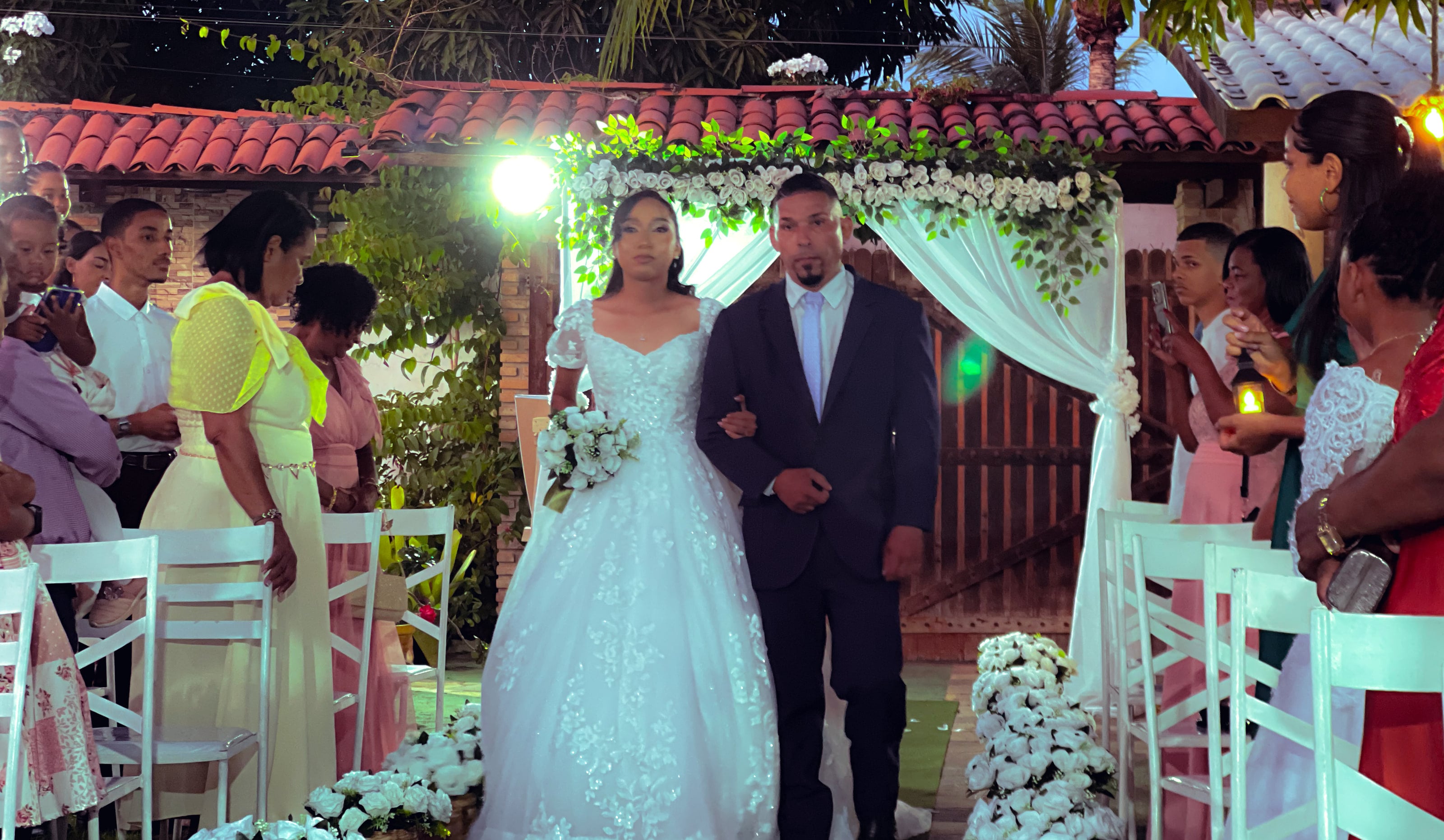 Entrada da noiva de braço dado pelo corredor de flores brancas, casamento de M & I