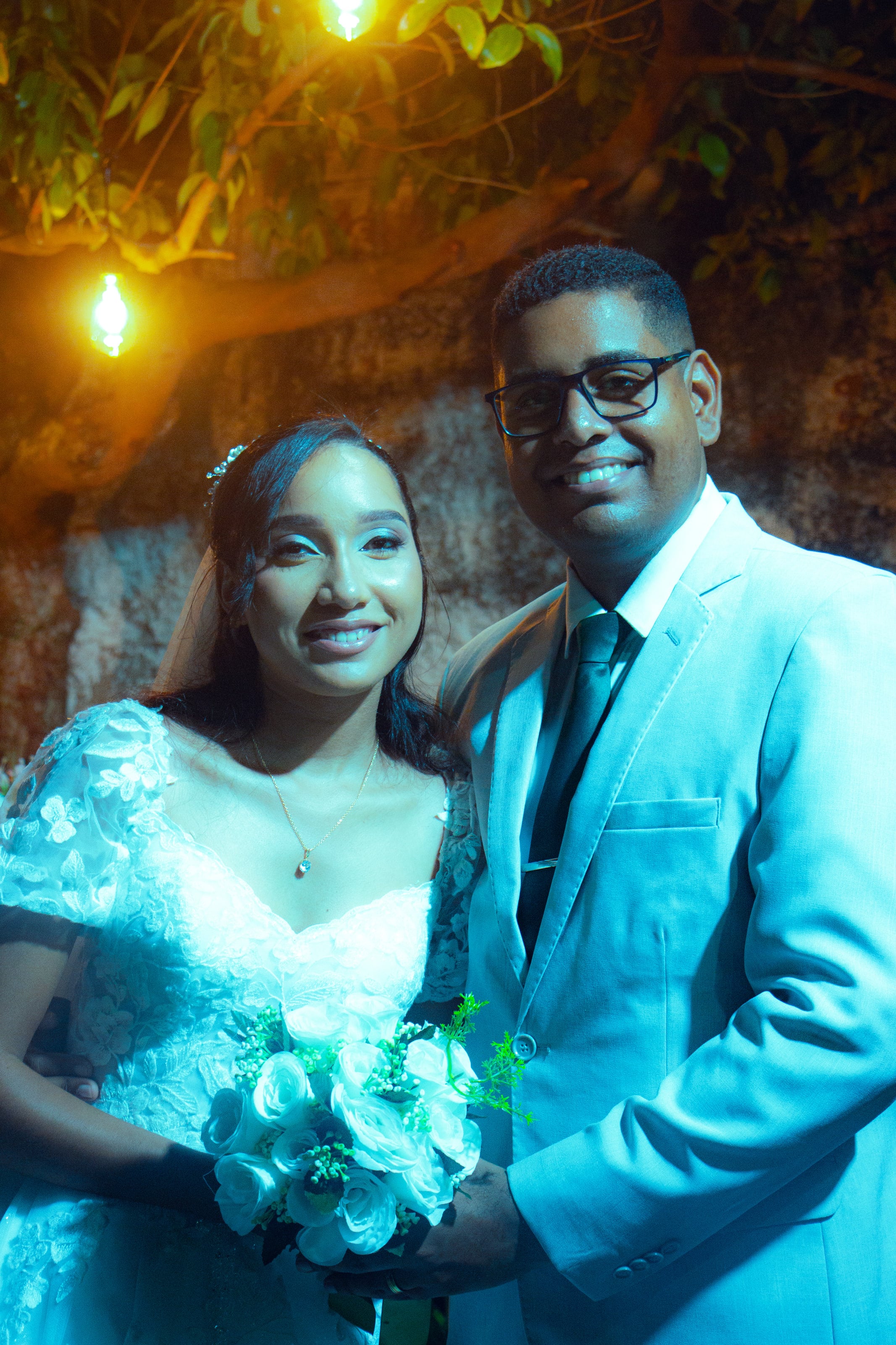 Casal junto à mesa do bolo, sob luzes quentes, casamento de M & I