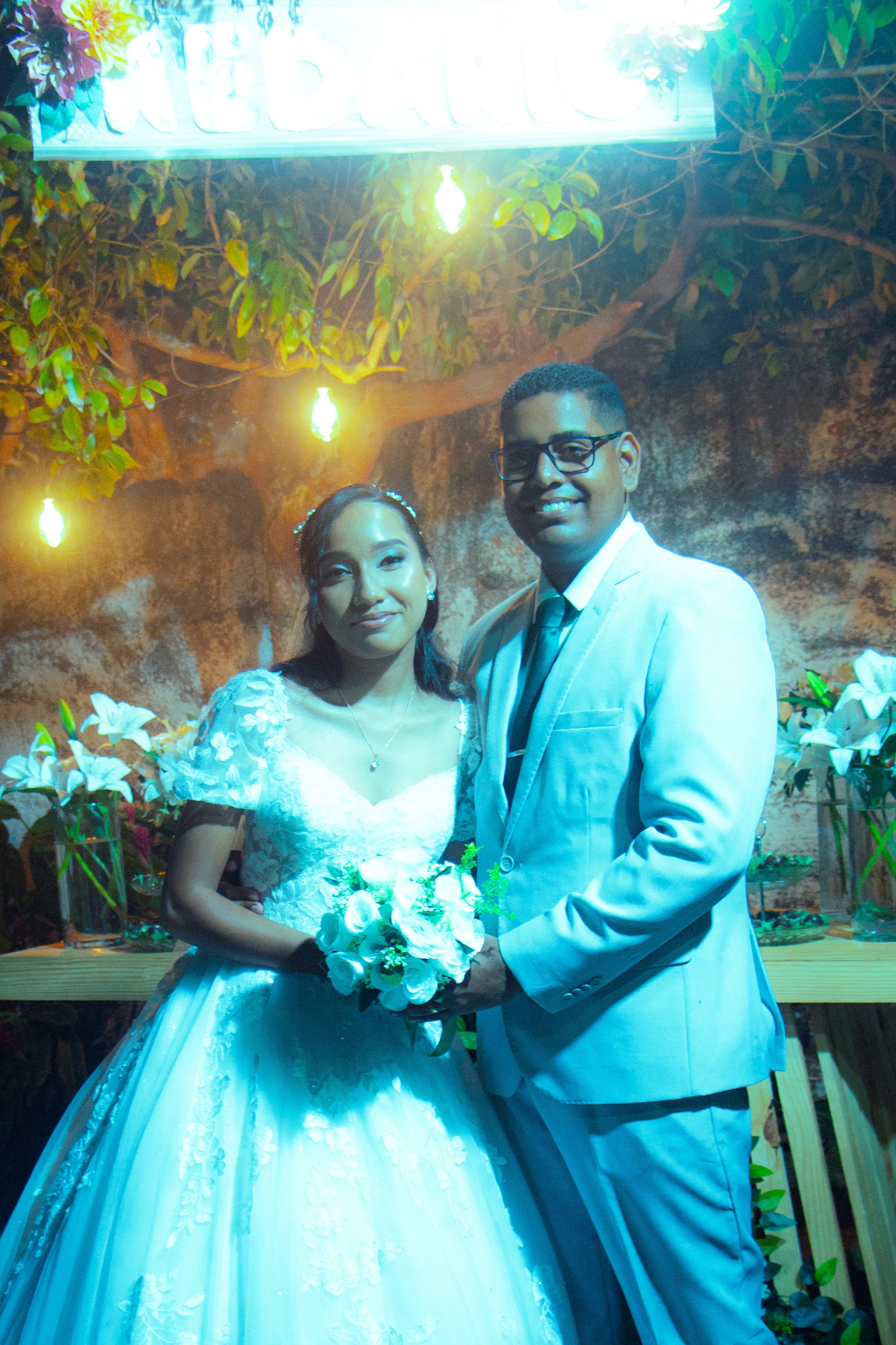 Casal junto à mesa do bolo, sob luzes quentes, casamento de M & I