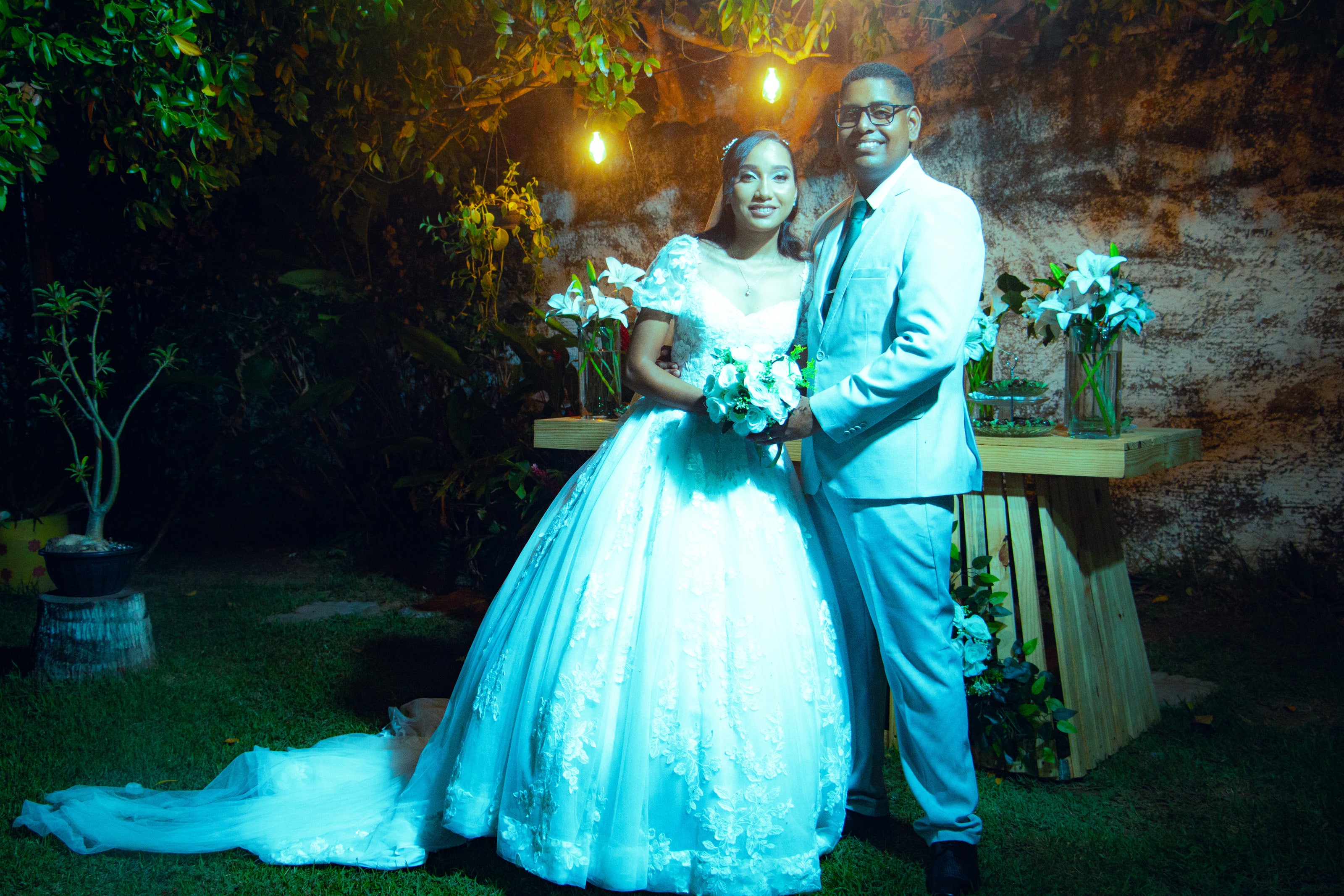 Casal junto à mesa do bolo, sob luzes quentes, casamento de M & I