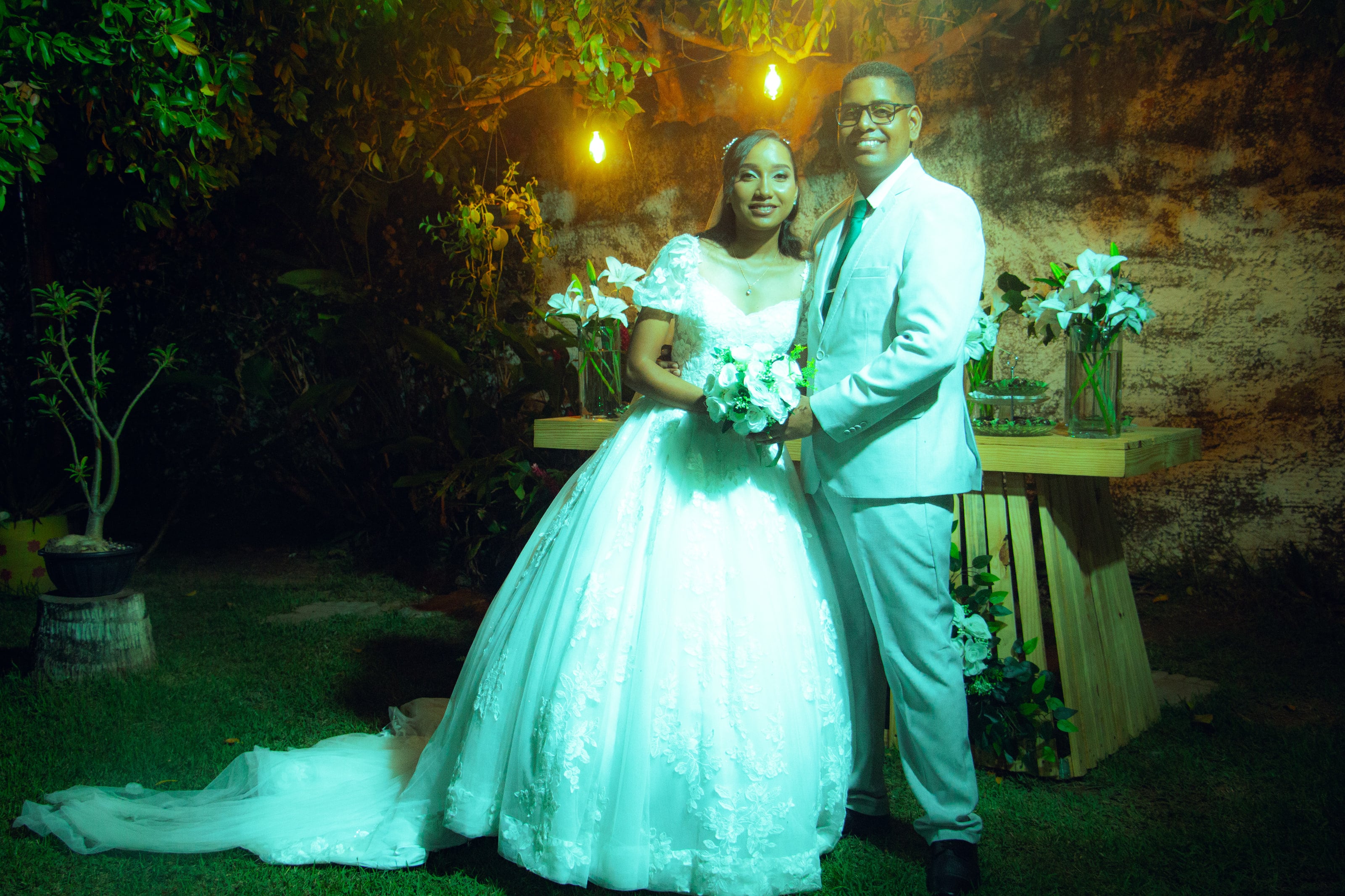 Casal junto à mesa do bolo, sob luzes quentes, casamento de M & I