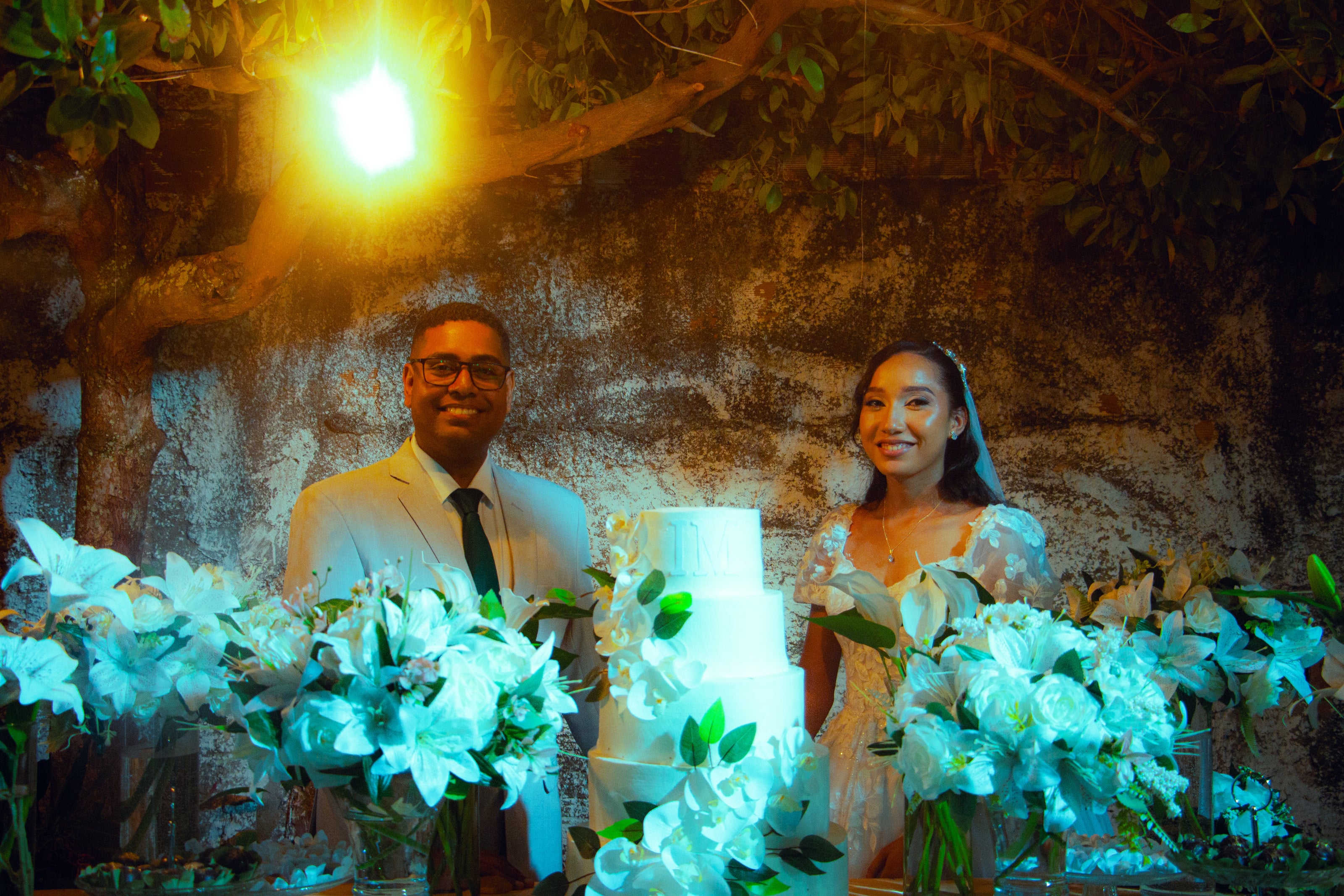 Casal junto à mesa do bolo, sob luzes quentes, casamento de M & I