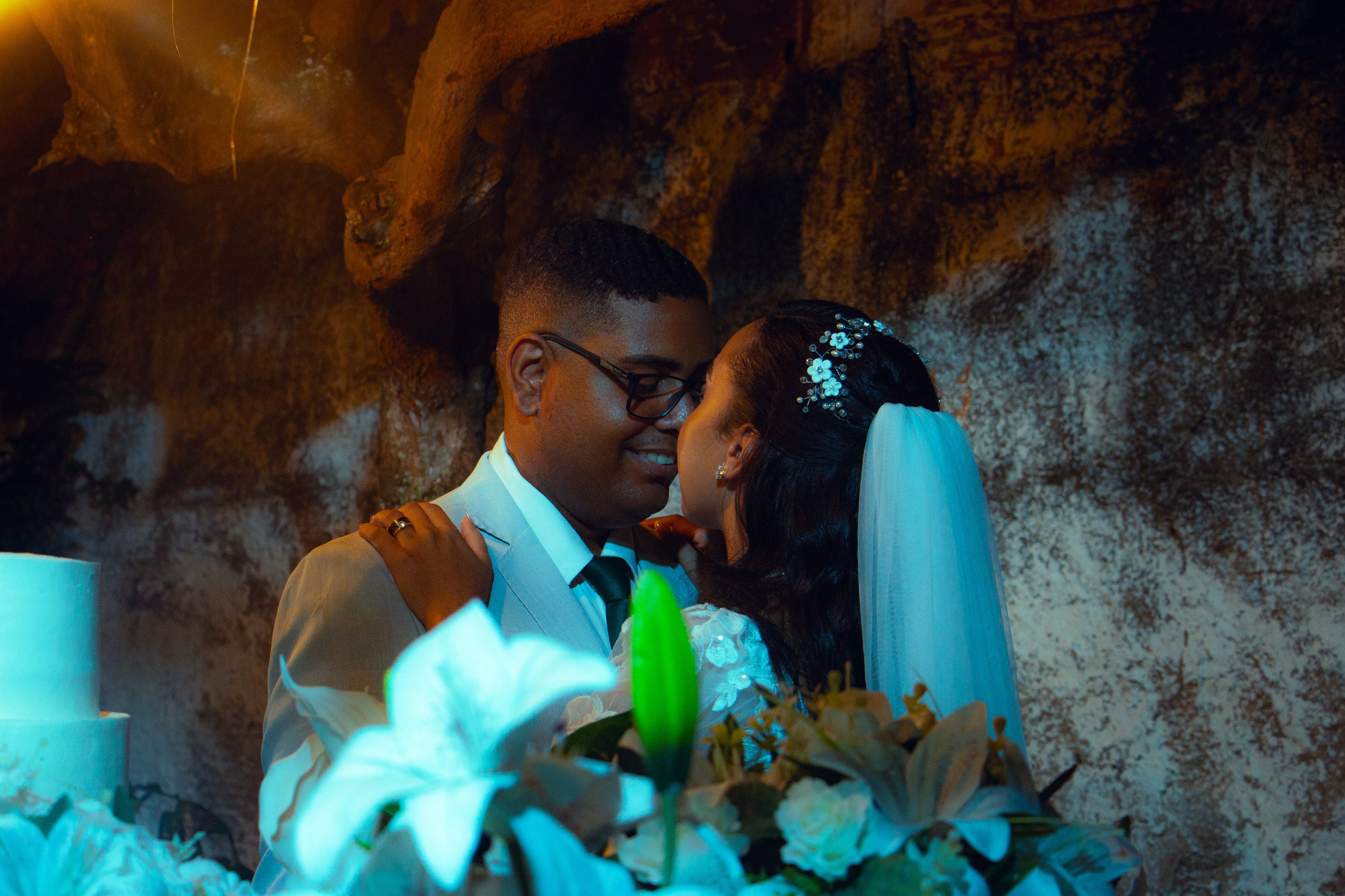 Casal junto à mesa do bolo, sob luzes quentes, casamento de M & I