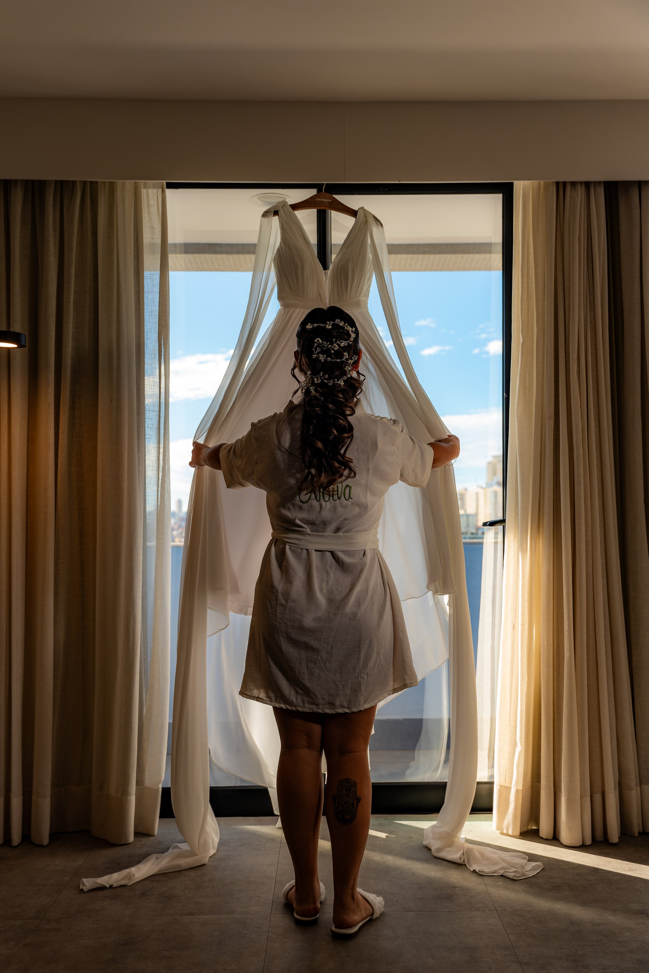 Noiva de costas segurando o vestido diante da janela, casamento de N & H