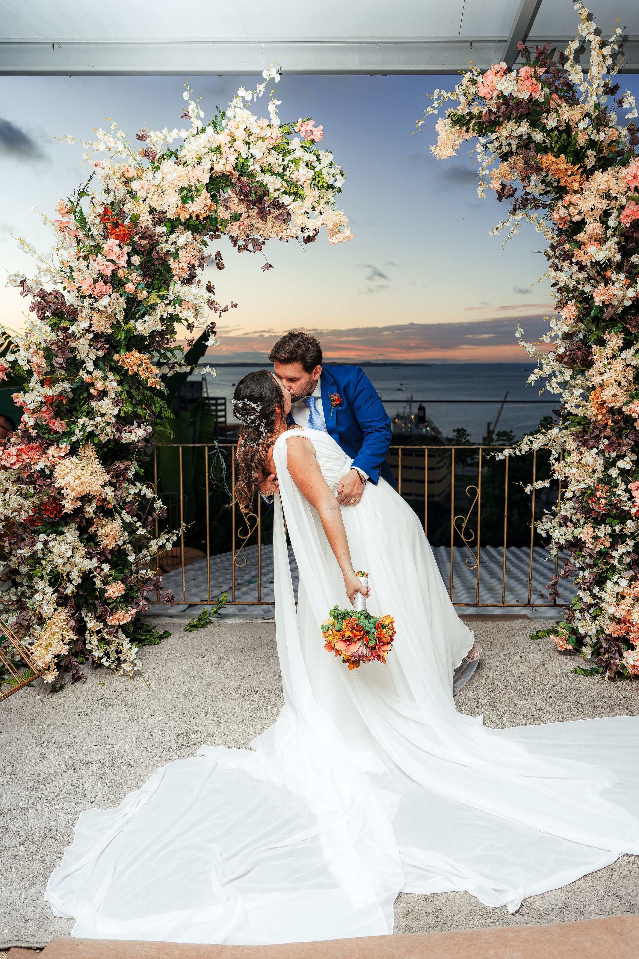 Beijo sob o arco de flores com o mar ao entardecer, casamento de N & H