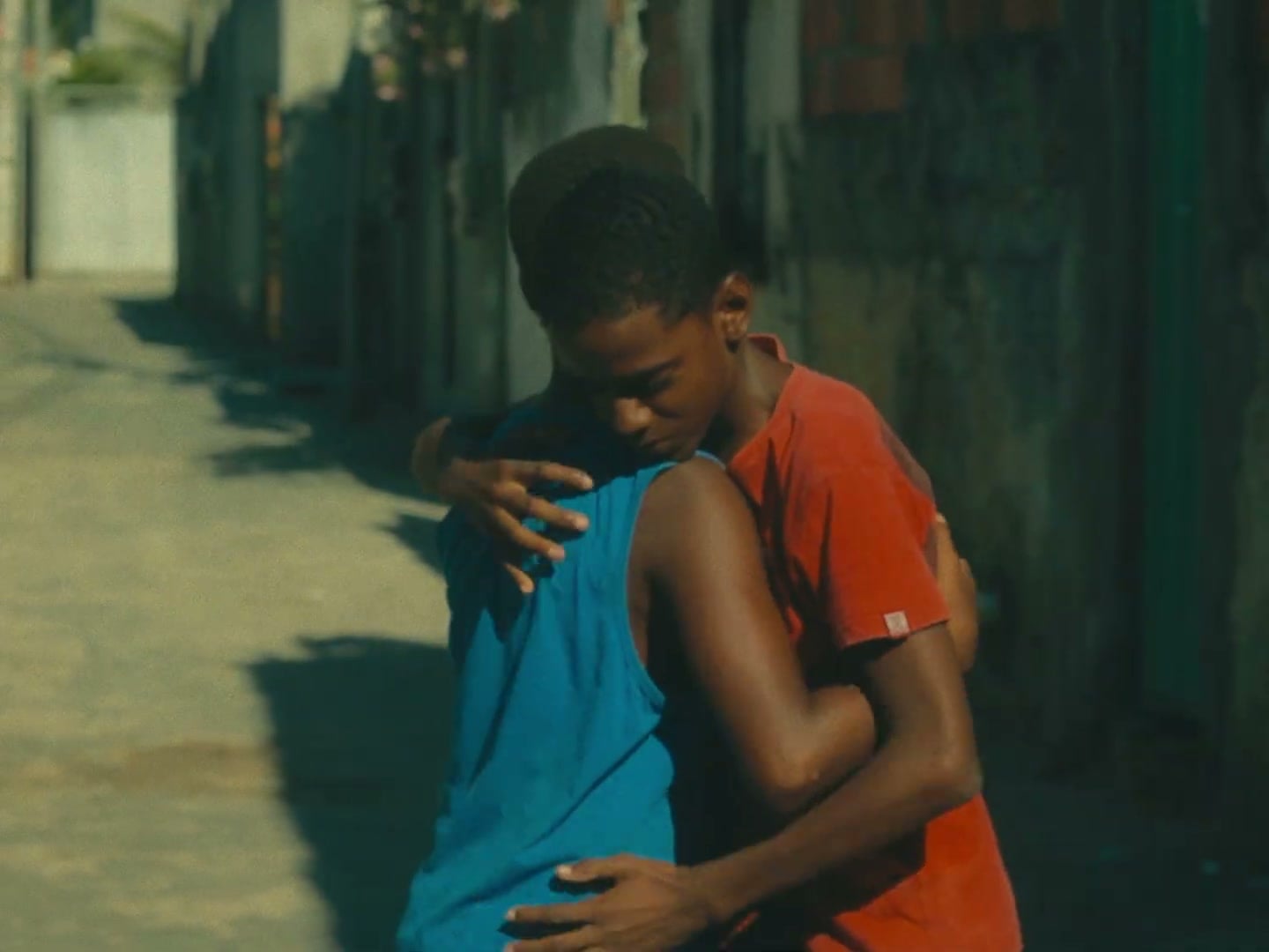 Dois adolescentes se abraçam no meio de uma rua estreita, sob sol forte.