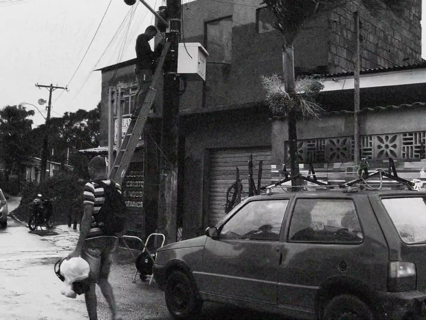 Rua de bairro em preto e branco, com um carro parado e caixas de som amarradas no bagageiro.
